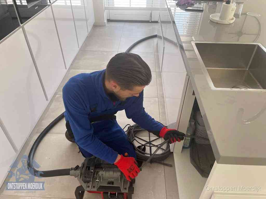 Een monteur ontstopt de keukenafvoer onder de spoelbak met een elektrische veermachine in Moerdijk