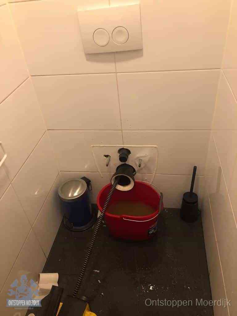 Een ontstopte wand-wc met verwijderde toiletpot waar een ontstoppingsveer in de wanduitgang verdwijnt, het achterhalen waar de verstopping zit in Moerdijk