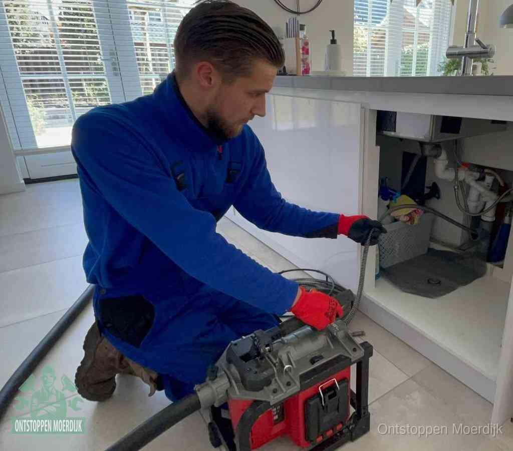 Een monteur met rode handschoenen knielt in een keuken en voert een elektrische ontstoppingsveer in tijdens het professioneel afvoer ontstoppen in Moerdijk