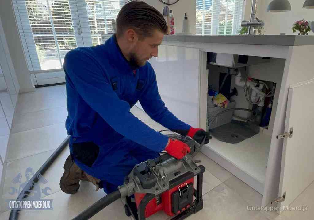 Een monteur voert een professionele elektrische ontstoppingsmachine in de afvoer onder de spoelbak tijdens het gootsteen ontstoppen in Moerdijk